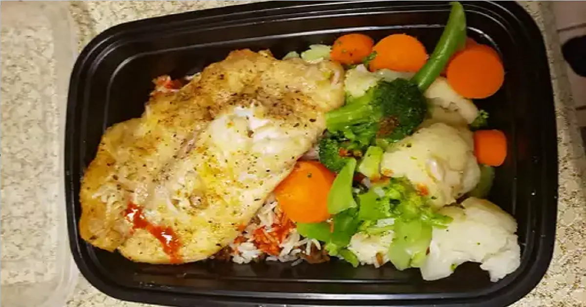 Pan-seared tilapia fillet on rice with steamed carrots, broccoli, cauliflower, and green beans in a black meal prep container.